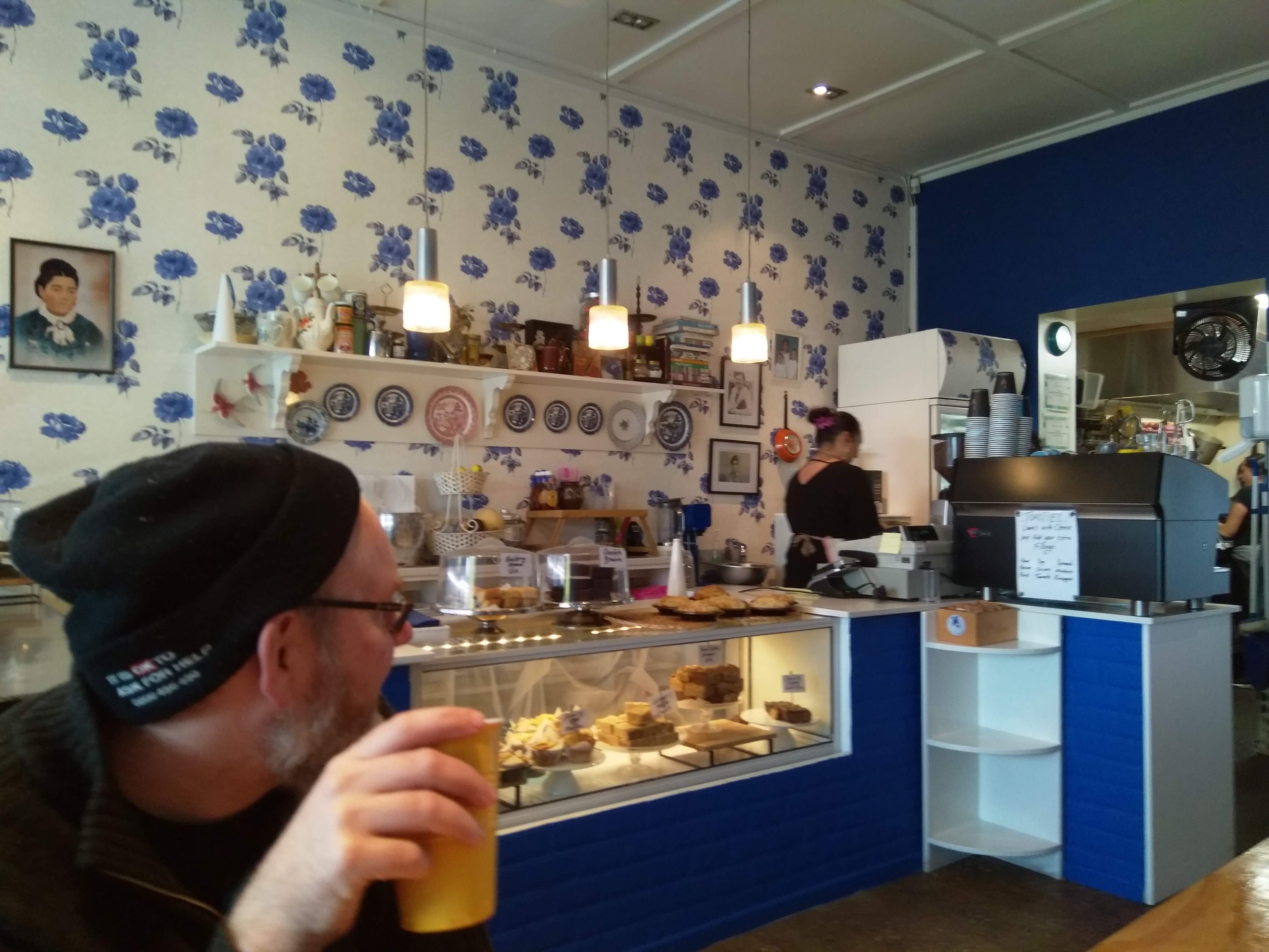 inside Blue Rose Cafe. Wallpaper behind the counter is white with blue roses. In the foreground a person sips coffee looking away from the camera.