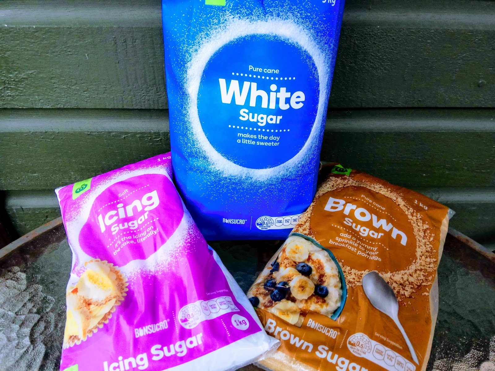 Three bags of sugar. A blue bag of white sugar leaning against a green wall. A pink bag of icing sugar and a brown bag of brown sugar lying along the flat surface.